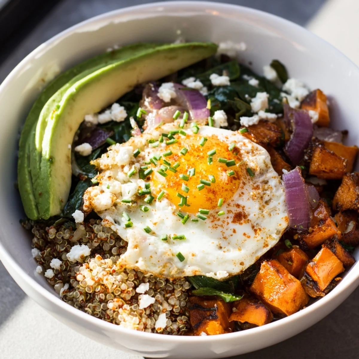 Hearty spinach and sweet potato quinoa breakfast bowl arranged in layers with creamy avocado slices and crumbled feta
