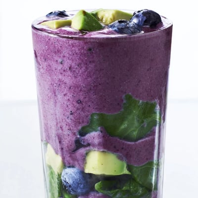 A chilled glass of Blueberry Avocado Spinach Smoothie with vibrant green and purple hues beside a halved avocado.